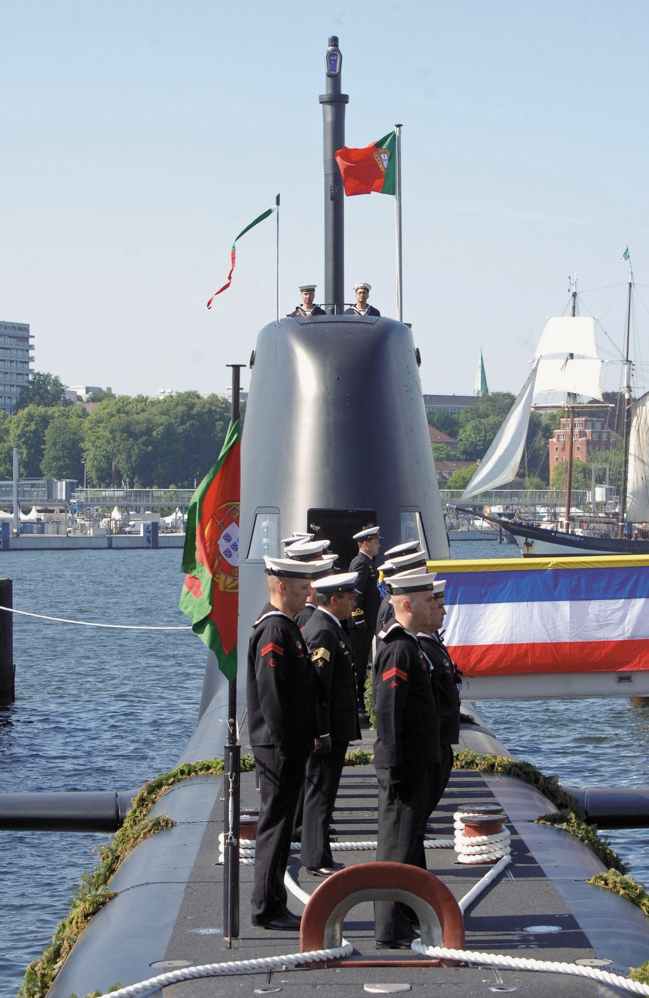 Portuguese navy personnel stand on a submarine deck during a ceremony with Portuguese flags displayed.