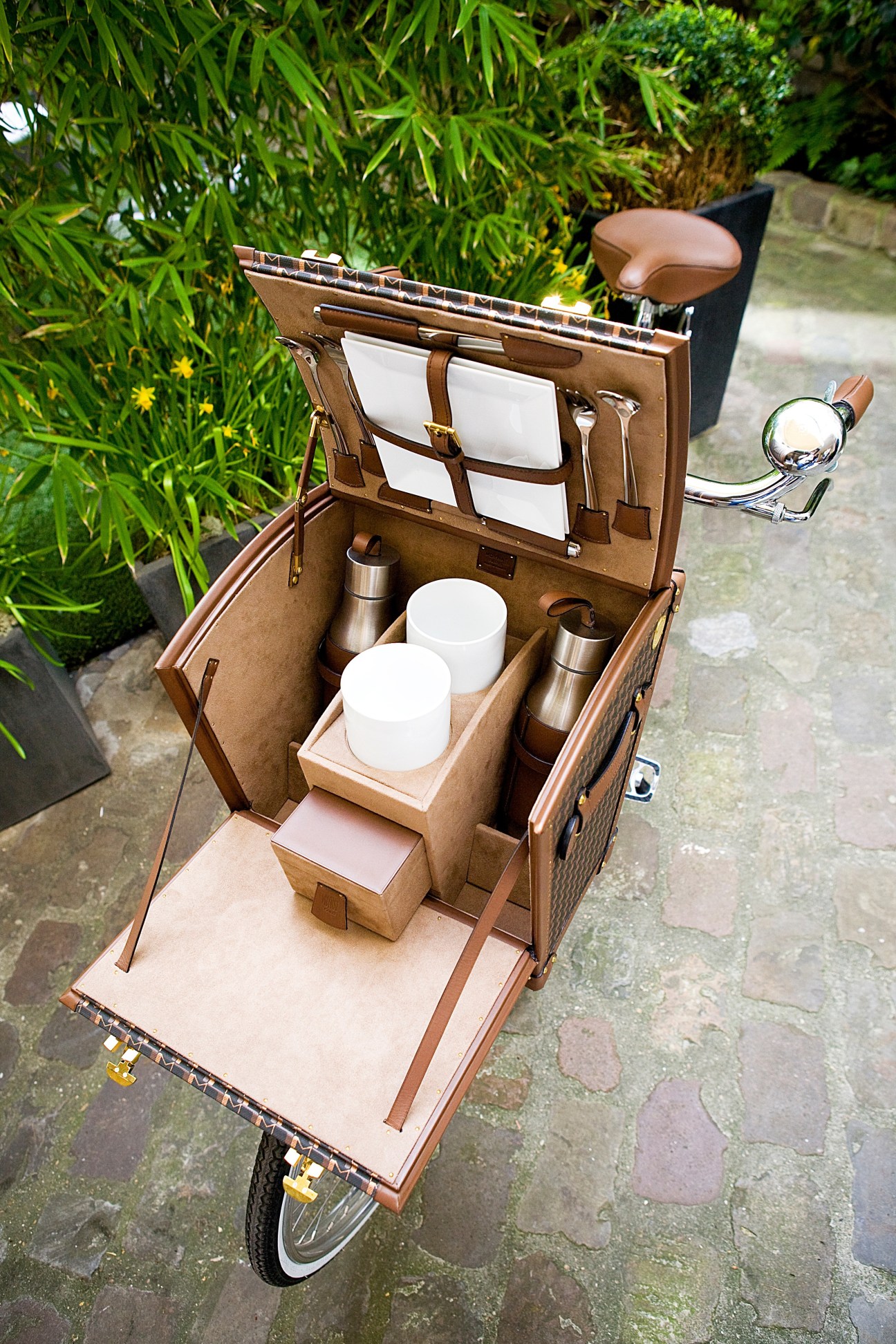 Moynat's Malle Picnic trunk open on a bicycle, displaying compartments with cups, containers and tableware.