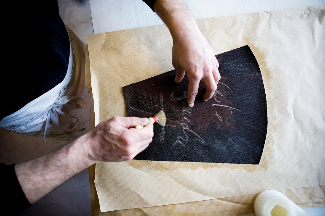 Hands applying glue to black monogrammed canvas for a Moynat trunk.