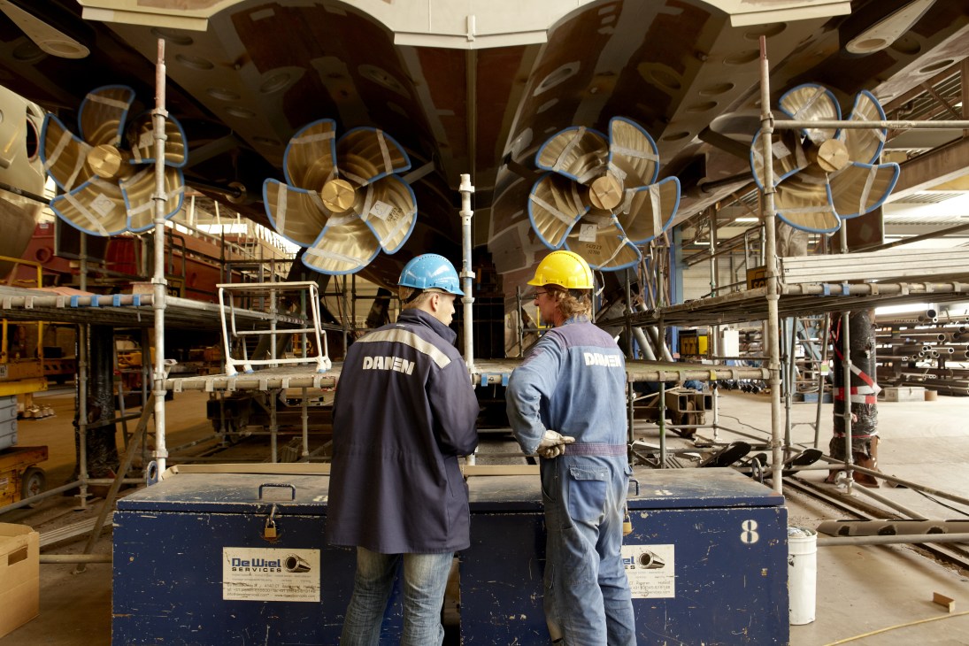 Two Damen shipyard workers in hard hats and branded clothing stand in an industrial factory space with large ship propellers and machinery.