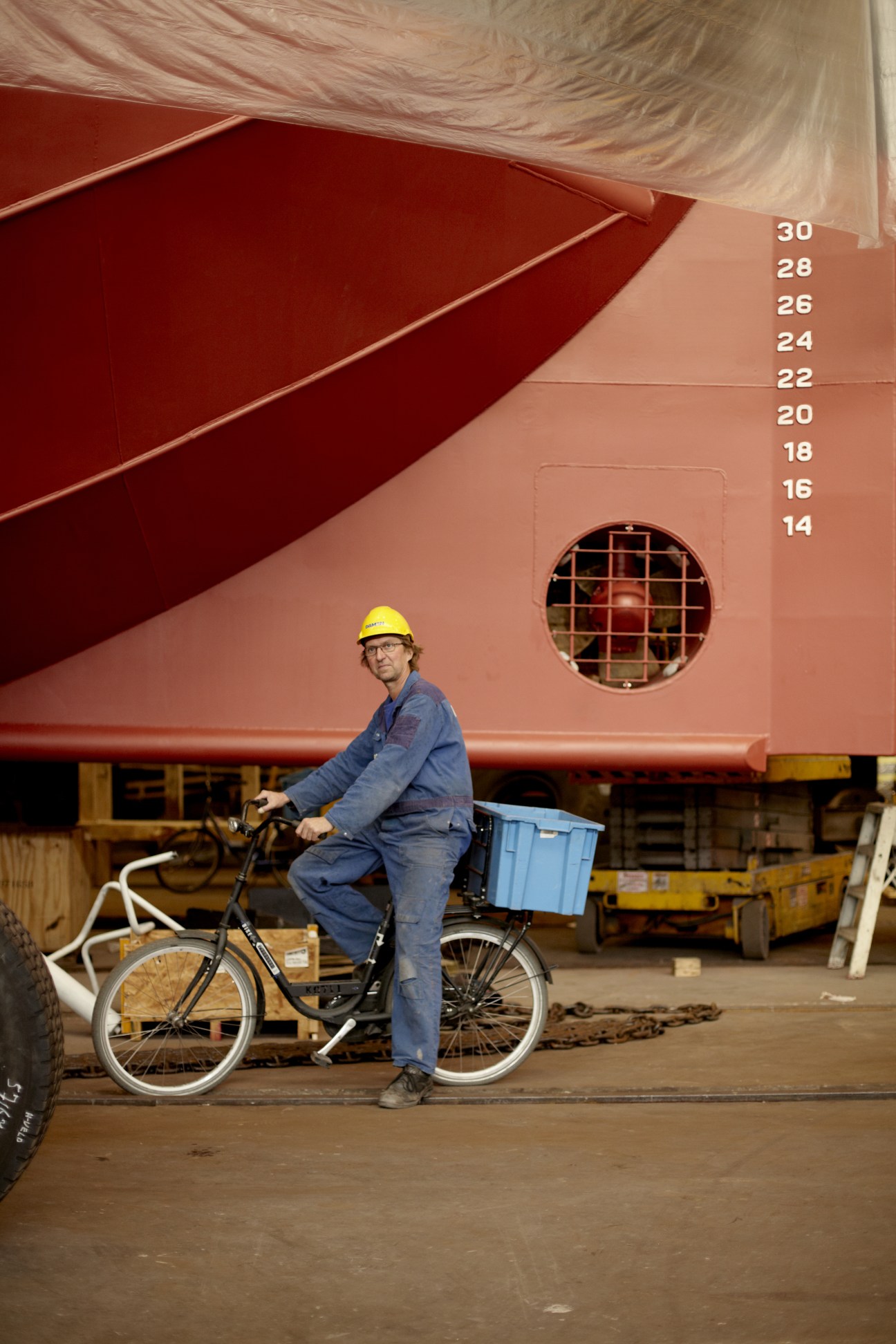 Worker on bicycle at Damen shipyard next to large red ship hull with measurement scale.