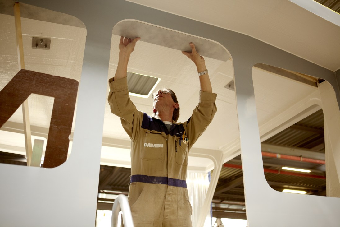 A carpenter in Damen work overalls installs a white curved panel inside a ship's bridge cabin.