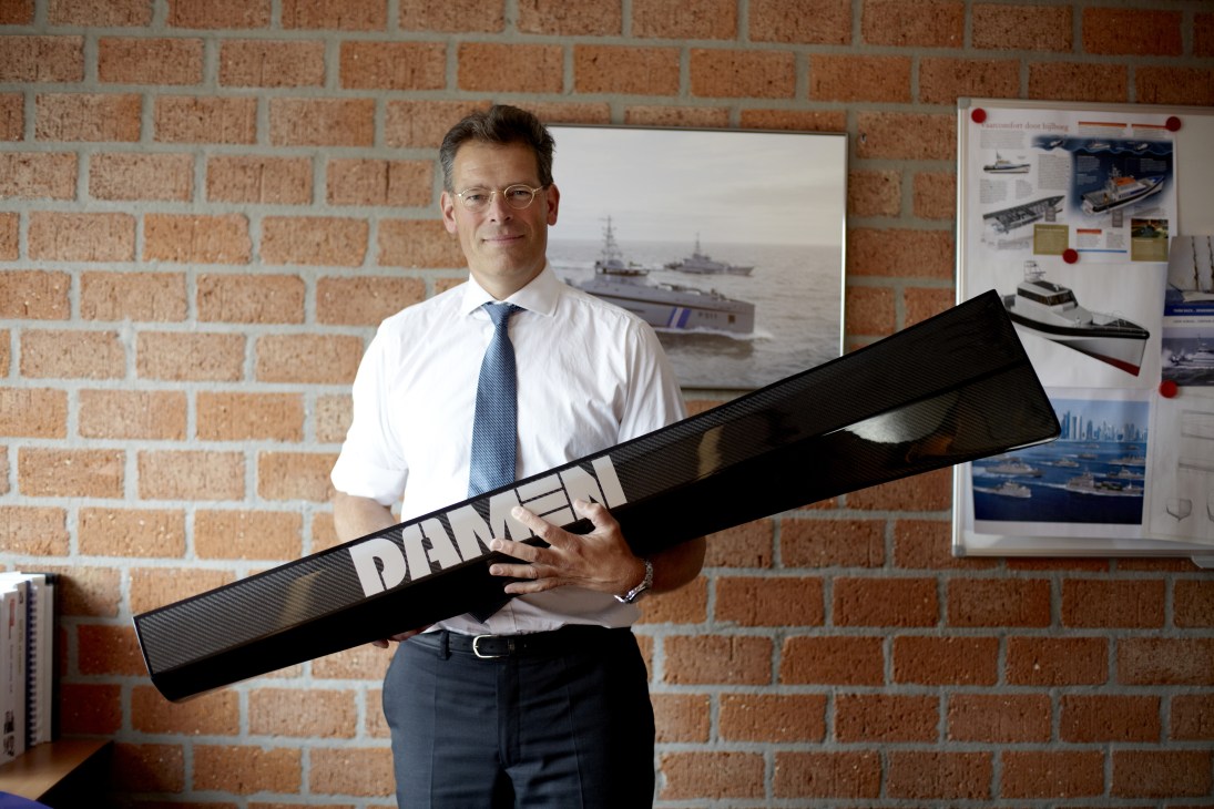 Jaap Gelling, product director of high-speed and naval craft, holds an oversized model of a ship's axe bow design in an office with exposed brick walls.