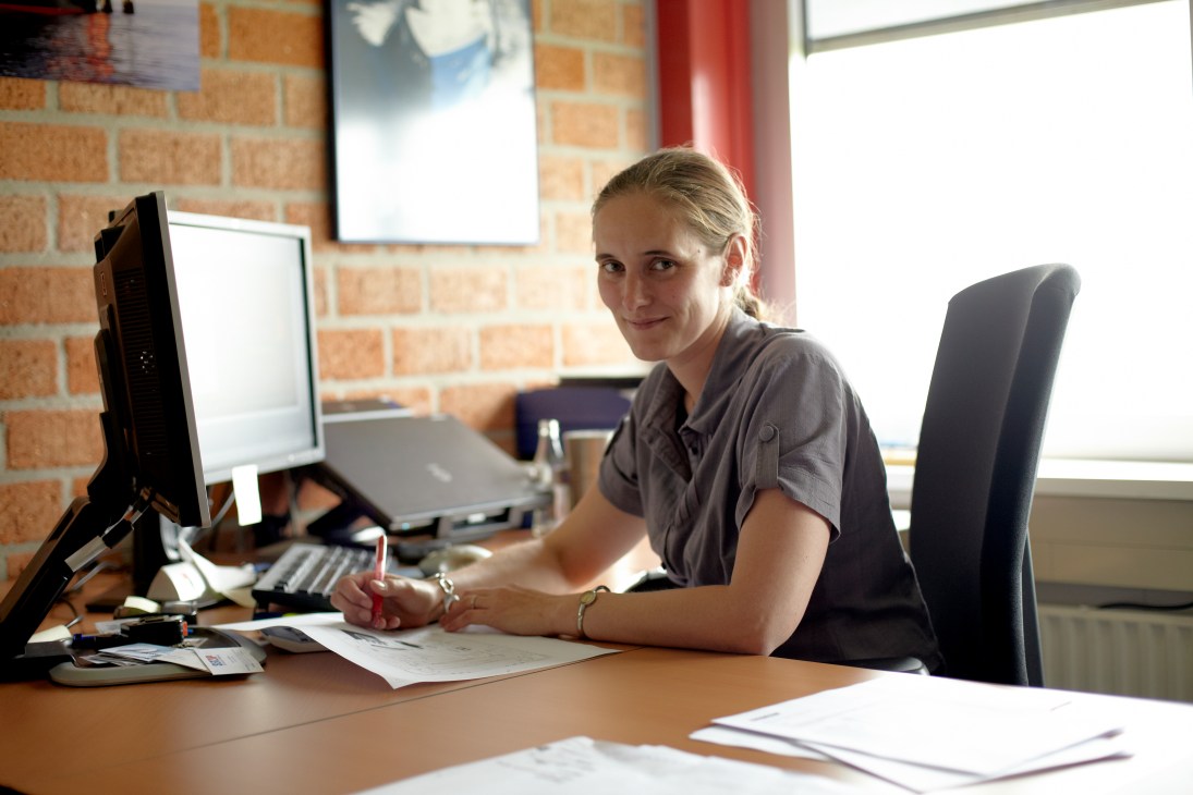 A woman in a grey shirt sits at a desk in a brick office, working on design drawings with a computer monitor nearby.