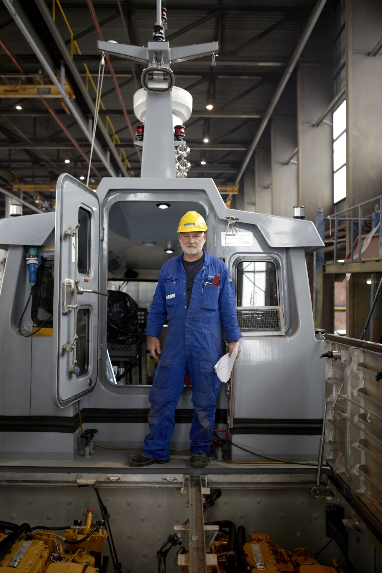 A foreman in blue overalls and yellow hard hat stands in the cabin of a patrol boat under construction at Damen shipyard.