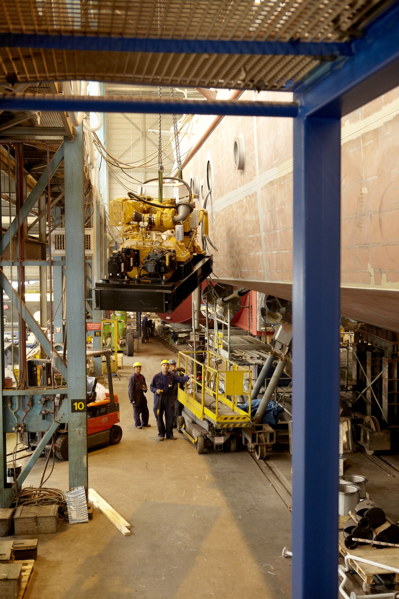 A yellow Caterpillar engine suspended by chain hoist above a ship hull in Damen shipyard.