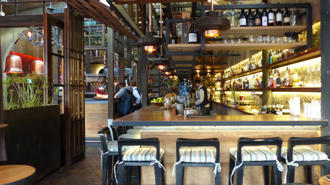 Interior of Woodpecker Hill restaurant showing bar seating with striped cushioned chairs, shelving with bottles, and warm lighting.