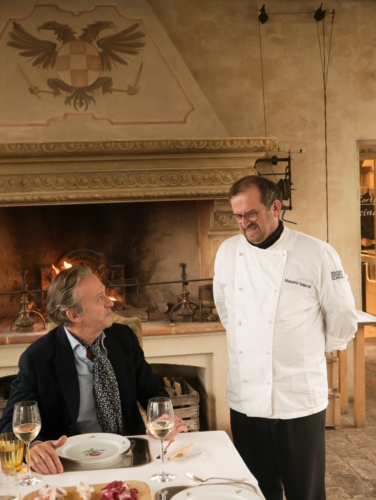 Umberto Angeloni sits at a dining table while chef Massimo Spigaroli stands beside him in a white chef's coat in a formal dining room with a fireplace.