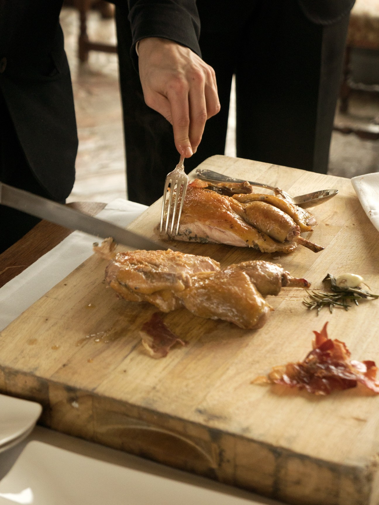 A hand with a fork cuts into roasted guinea fowl on a wooden cutting board during plating.