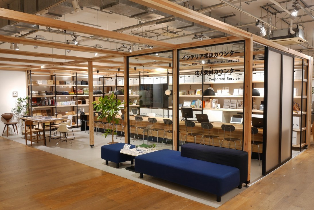 Modern interior consultation space with wooden frame structure, glass partitions, blue seating, and shelving displaying Muji products and materials.