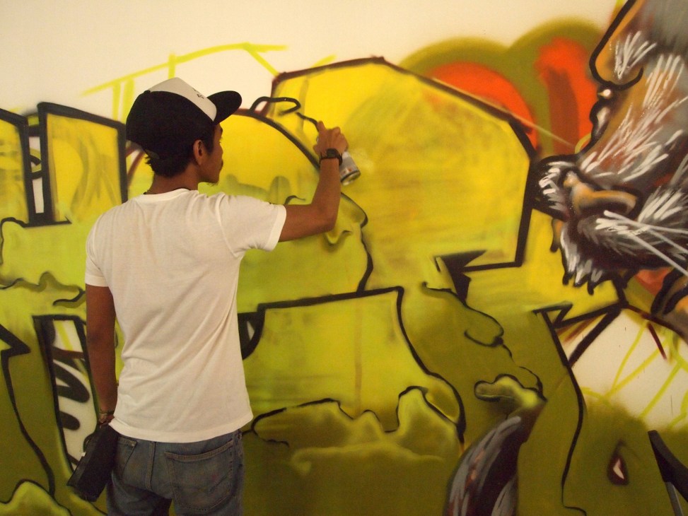 Person in white shirt and cap spray-painting yellow and black graffiti art on a wall.
