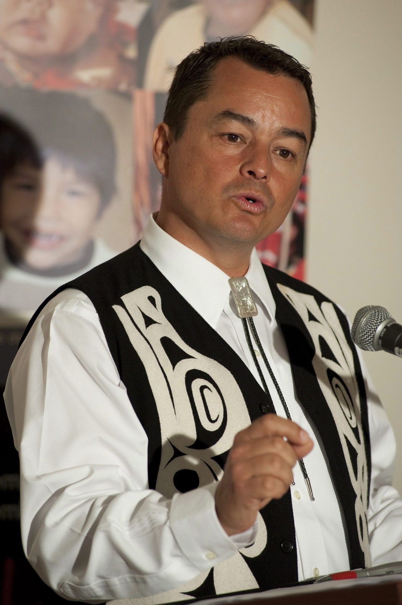 Indigenous leader in ceremonial regalia speaking at a microphone during an assembly.