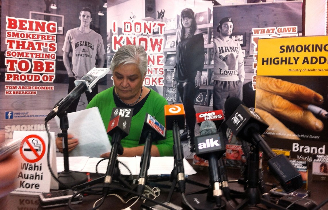 Woman at press conference speaking about tobacco control policy with anti-smoking campaign materials visible behind her.