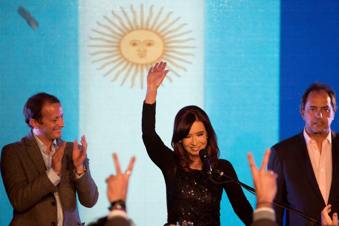 Woman at podium raises fist while two men applaud during Argentine election campaign event.