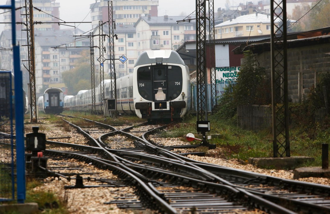 Modern Talgo high-speed train parked unused on a Sarajevo railway siding surrounded by overgrown tracks and electrical infrastructure.