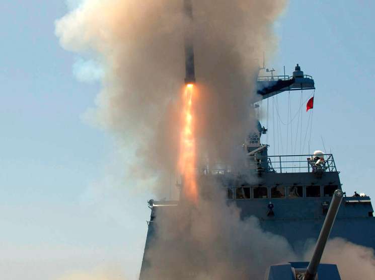 A naval vessel fires a cruise missile into the sky amid smoke and flame.