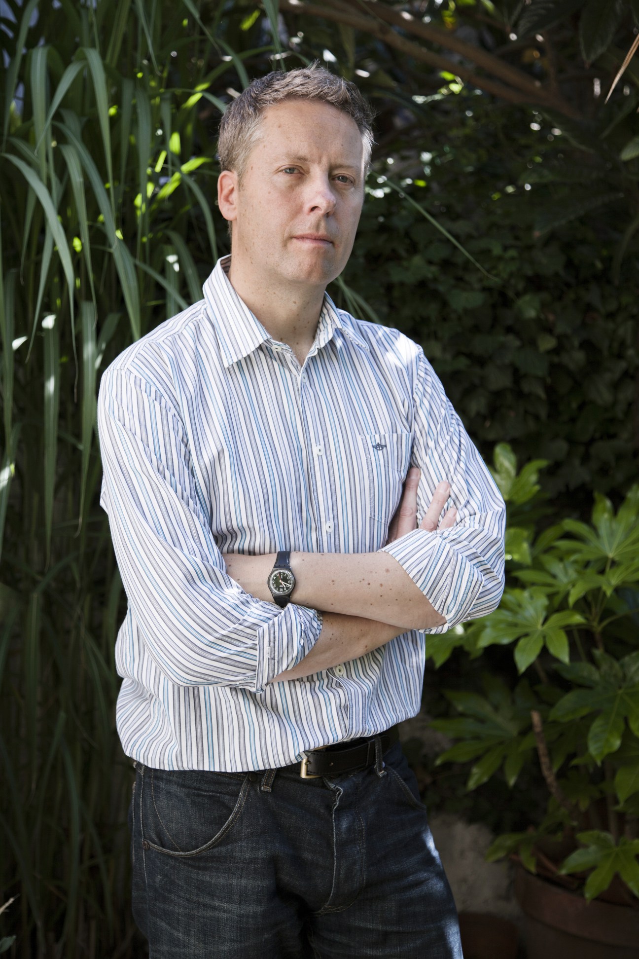 Matthew Cook, a war artist, poses with arms crossed wearing a blue striped shirt against a leafy green background.