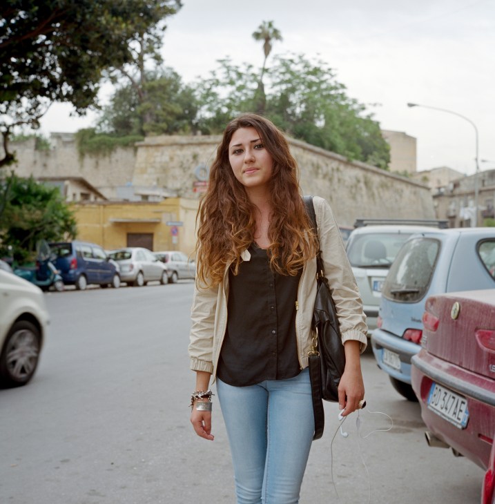 Young woman with long reddish-brown hair standing on a Palermo street lined with parked cars and historic buildings.