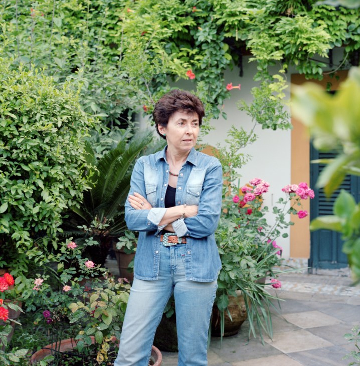 Nicoletta Polo, Duchess of Palma, stands with crossed arms in a lush garden surrounded by flowering plants and climbing ivy.