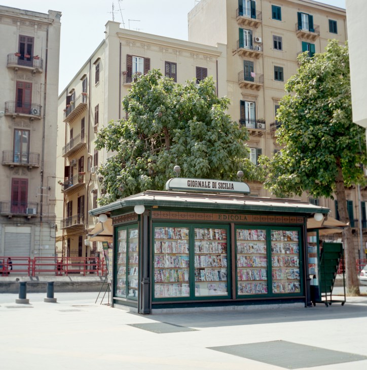A green newsstand kiosk with displayed magazines surrounded by ivy, in front of historic Palermo buildings.