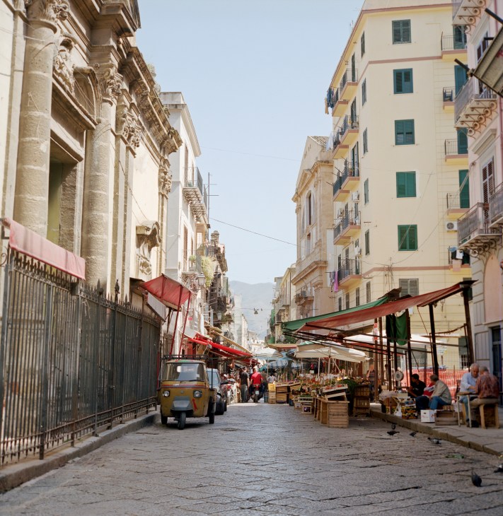 A bustling street market in Via Porta Carini, Palermo, with colorful awnings, vintage vehicles, and historic architecture lining both sides.