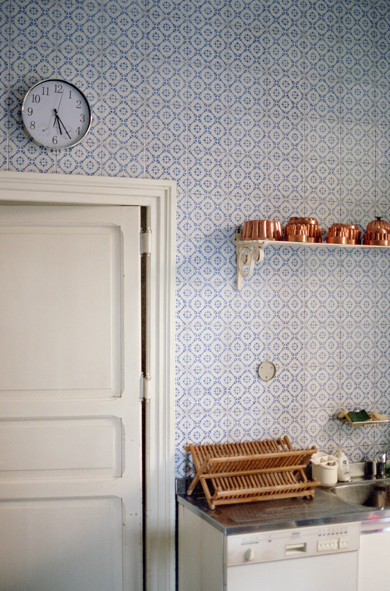 Kitchen with blue patterned wallpaper, white door, wall clock, copper cookware on shelf, and wooden dish rack on counter.