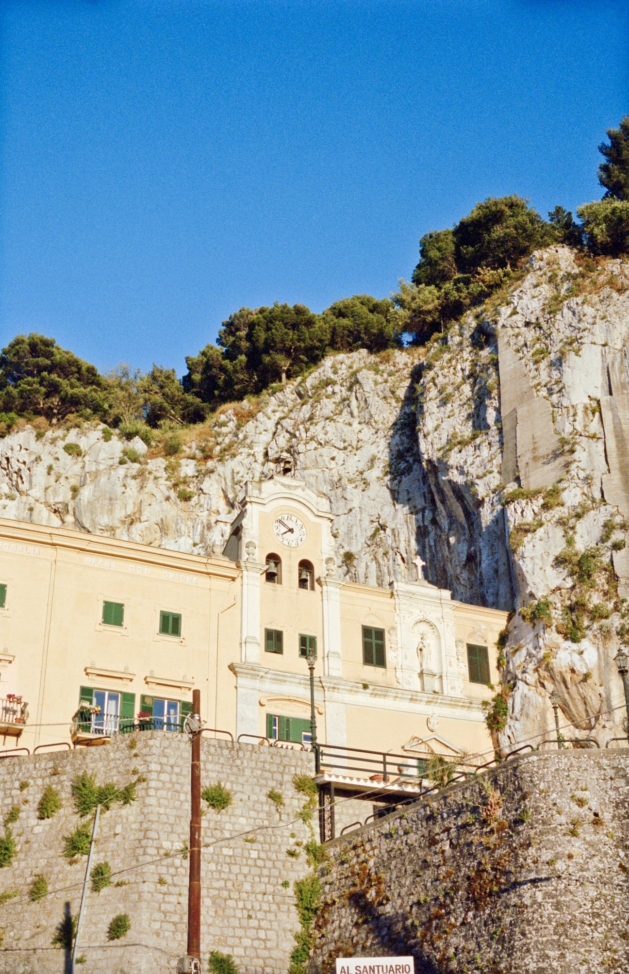 Sanctuary of Santa Rosalia built into a limestone cliff with cream-colored facade and green shutters.