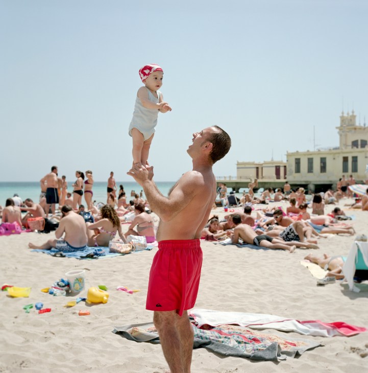 A man lifts a toddler wearing a pink and white hat on a crowded beach at Mondello, with a colonial-style building visible in the background.