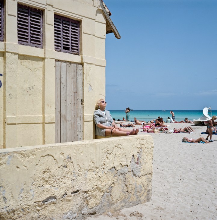 Woman sitting against a cream-colored building with shuttered windows, relaxing on Mondello beach with beachgoers and turquoise sea visible beyond.