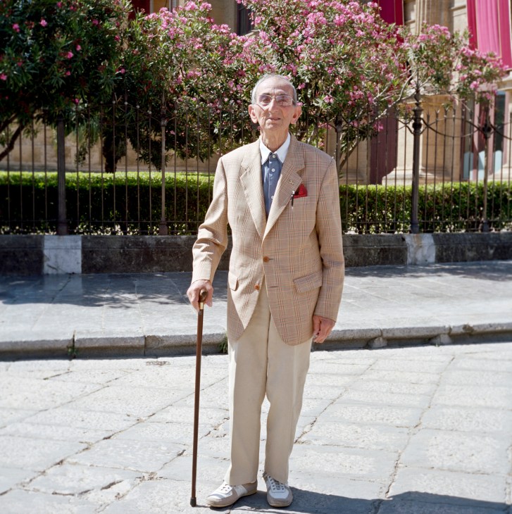 Elderly man in beige suit with cane stands on Via Re Federico in Palermo, pink flowering plants visible behind iron fence.