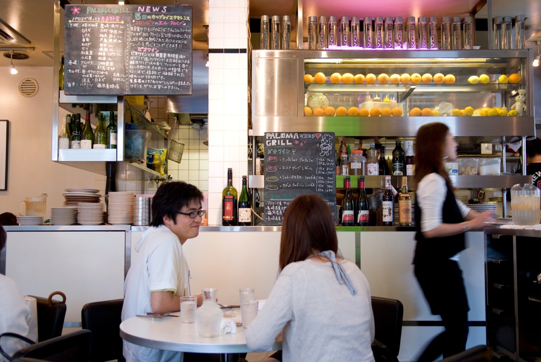 Interior of Paloma Grill restaurant with customers seated at a table and staff behind counter displaying fresh citrus fruits and beverages.