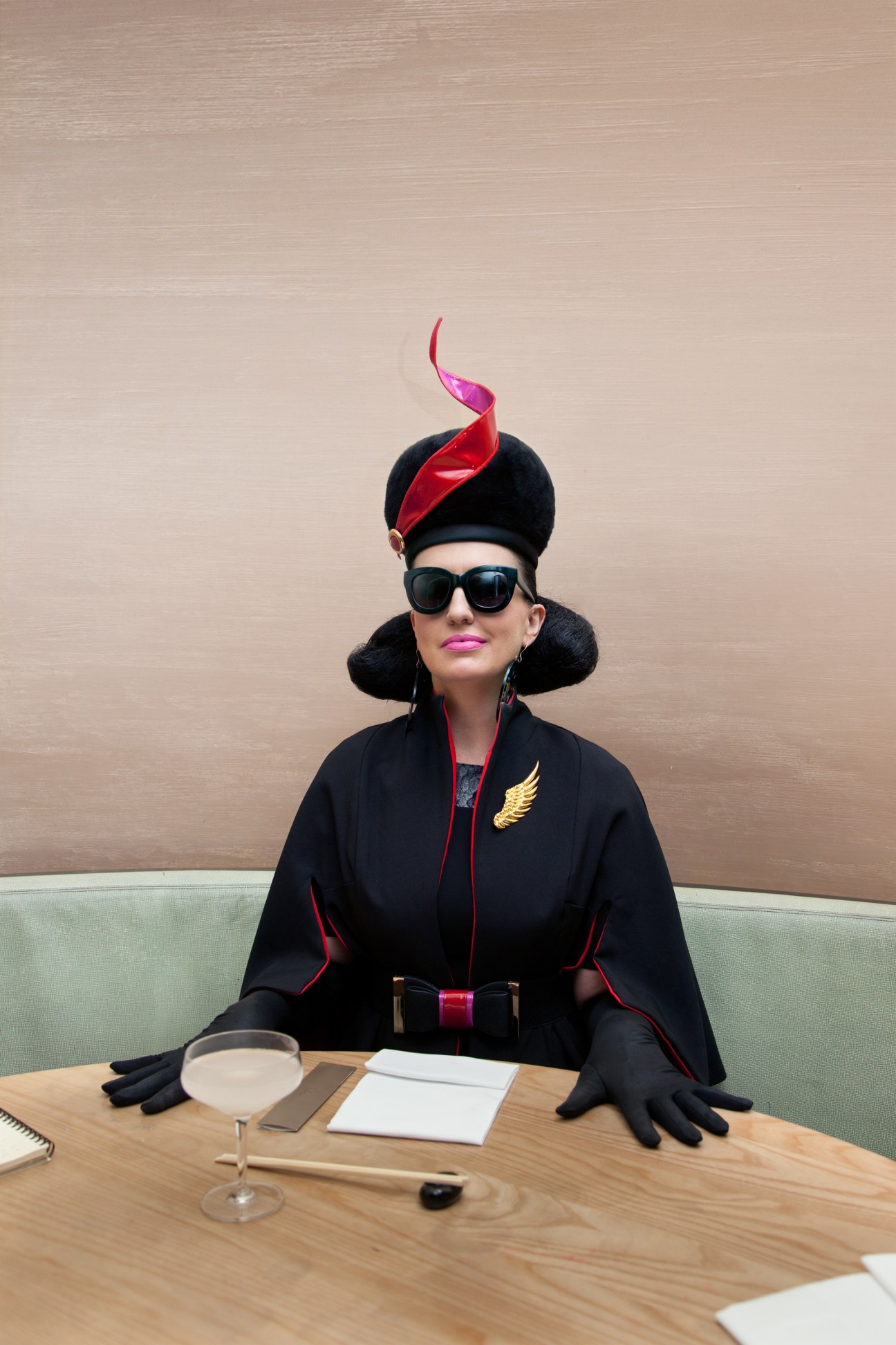 Pam Ann character in black uniform with red and gold accents, oversized black hat with red plume, and sunglasses, seated at a table.