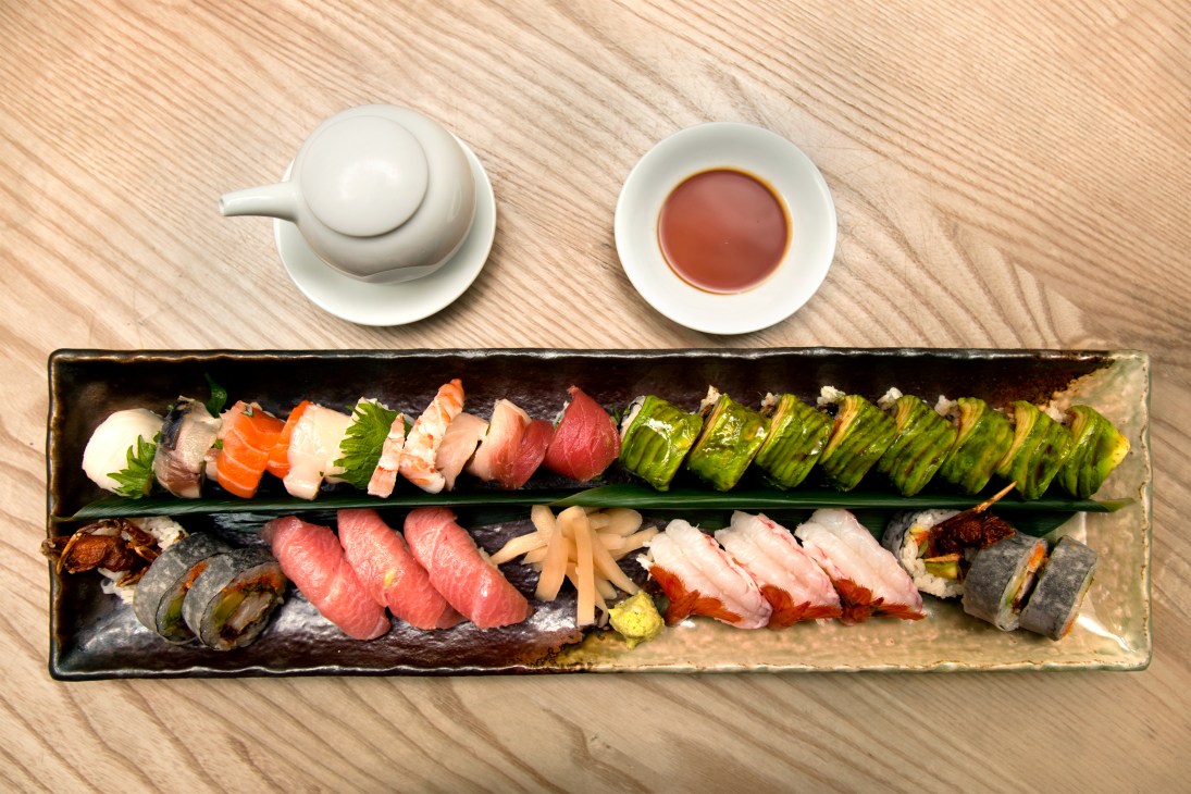 Assorted sushi and sashimi arranged on a dark rectangular plate with white condiment bowls on wooden table.