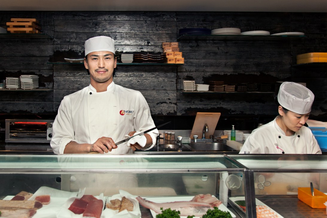 Two Nobu chefs in white uniforms and headbands working behind the sushi counter with ingredients displayed below.