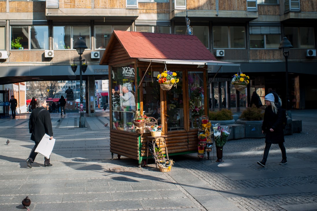 A wooden flower kiosk with a red roof on a Belgrade pedestrian street, displaying colorful flowers in baskets.