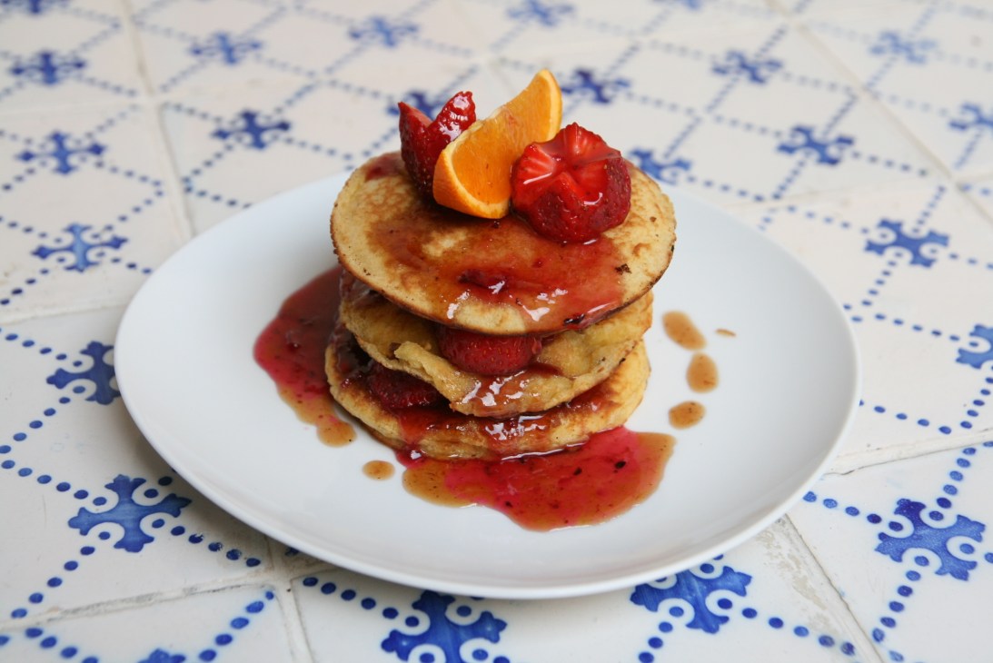 Stack of three pancakes topped with strawberries, orange slice and red sauce on white plate.