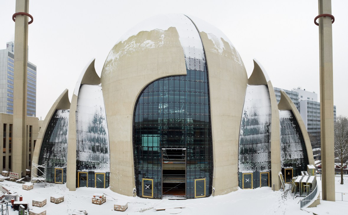 Central Mosque in Köln under construction, covered in snow, featuring distinctive pale concrete dome and curved architectural elements.