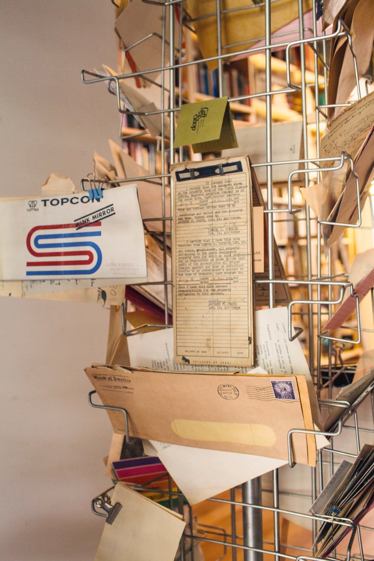 Metal wire shelving unit displaying vintage books, magazines, envelopes and ephemera including a Topcon Pink Mirror publication.