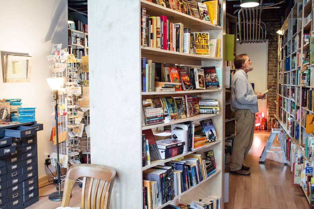 Interior of The Paper Hound bookstore showing white shelving units filled with colorful books and a man browsing.