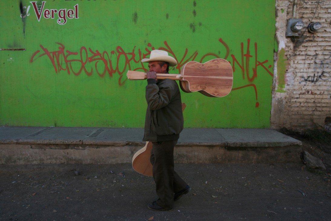 A man in a cowboy hat carries a wooden classical guitar over his shoulder past a green wall with graffiti.