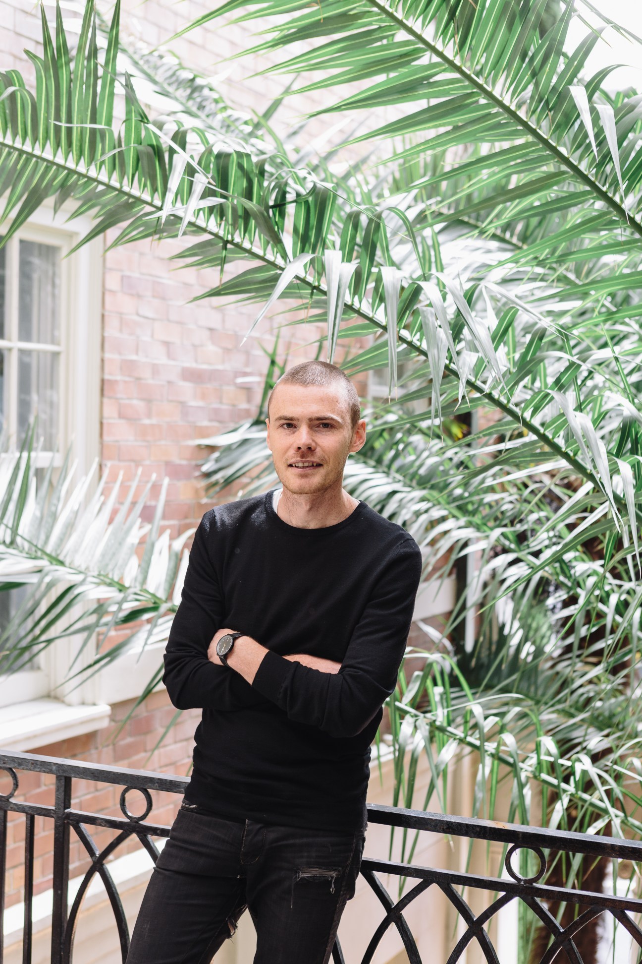 Interior designer Rufus Knight stands on a brick balcony beneath hanging green palm fronds.