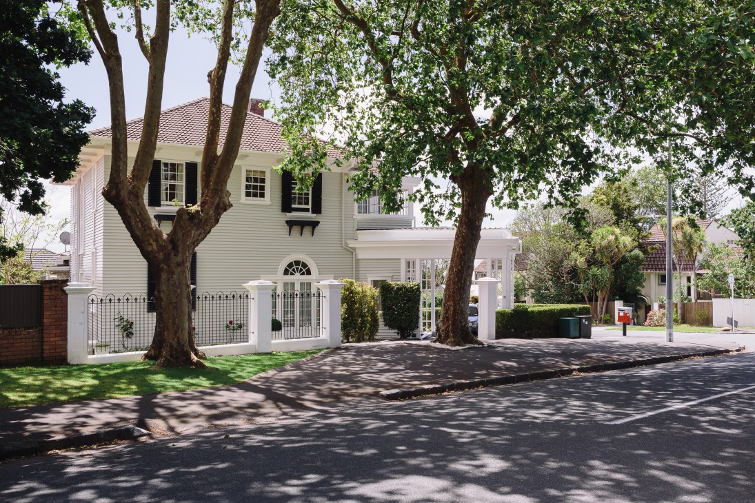 White wooden villa with black shutters shaded by large trees on a quiet Parnell street.