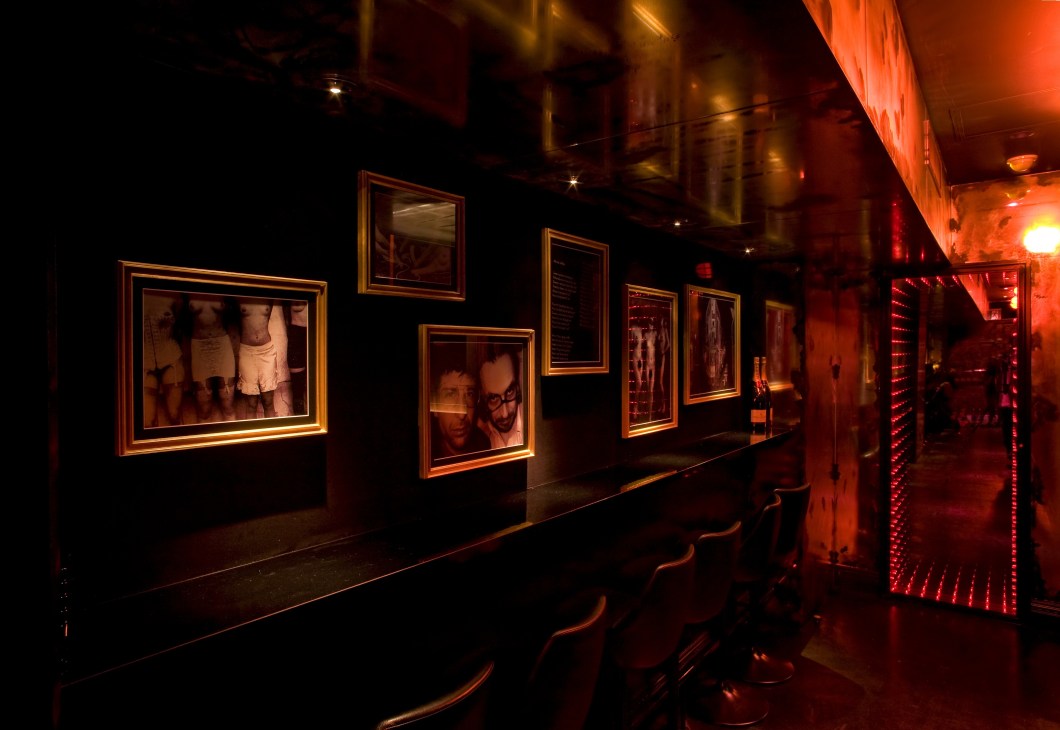 A dimly lit corridor with black walls lined with framed black-and-white photos and sparkling mirrors, illuminated by warm red and gold lighting.