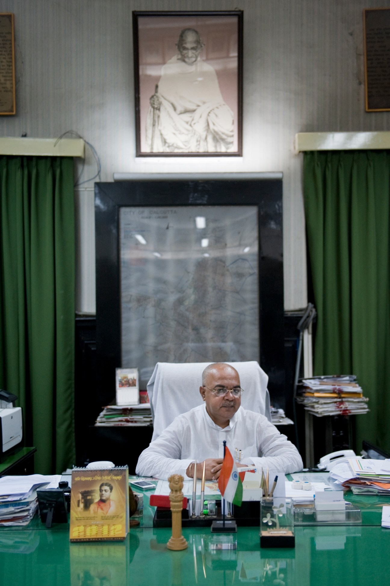 Mayor Sovan Chatterjee seated at his desk in an official office with a portrait of Gandhi hanging on the wall above.