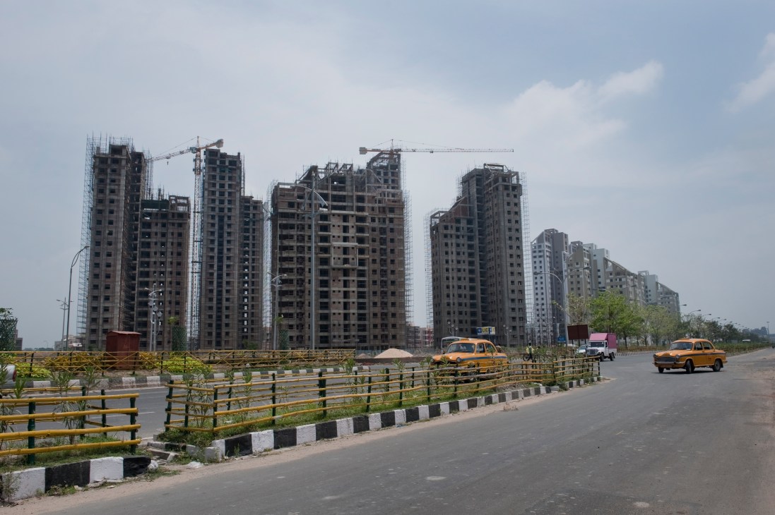 Construction cranes tower over multi-story residential buildings under development in Rajarhat, Kolkata, with yellow taxis on an empty road.