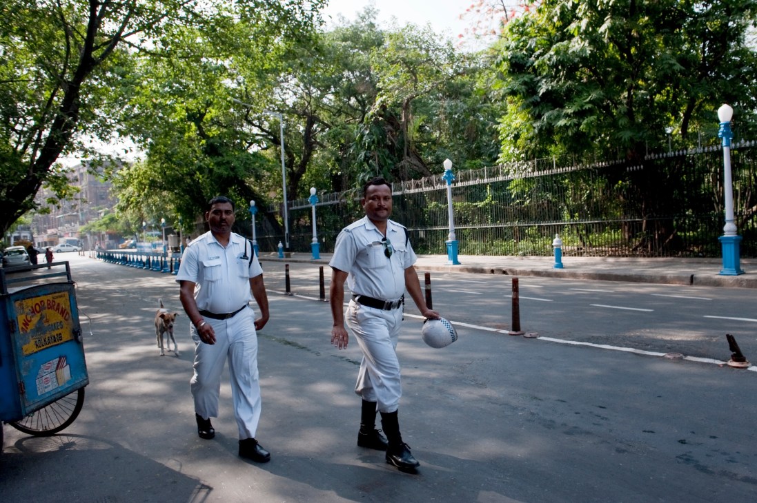 Two police officers in white uniforms patrol a tree-lined street in Kolkata near Raj Bhavan.