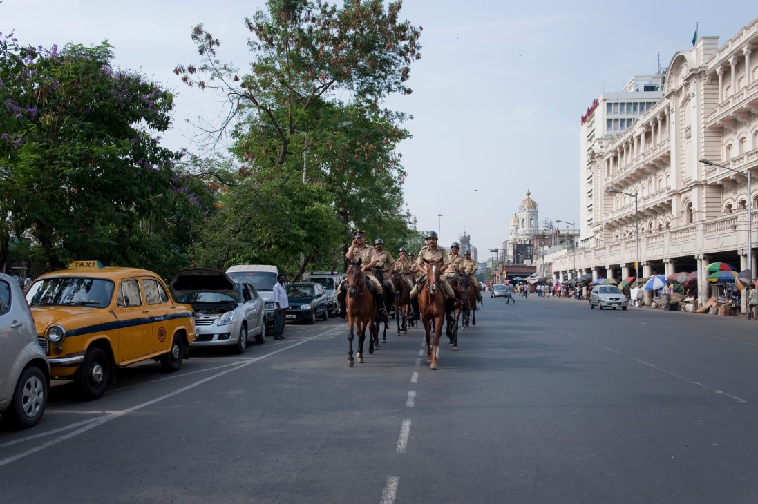 Police officers on horseback patrol a wide tree-lined street in Kolkata past colonial architecture and yellow taxis.