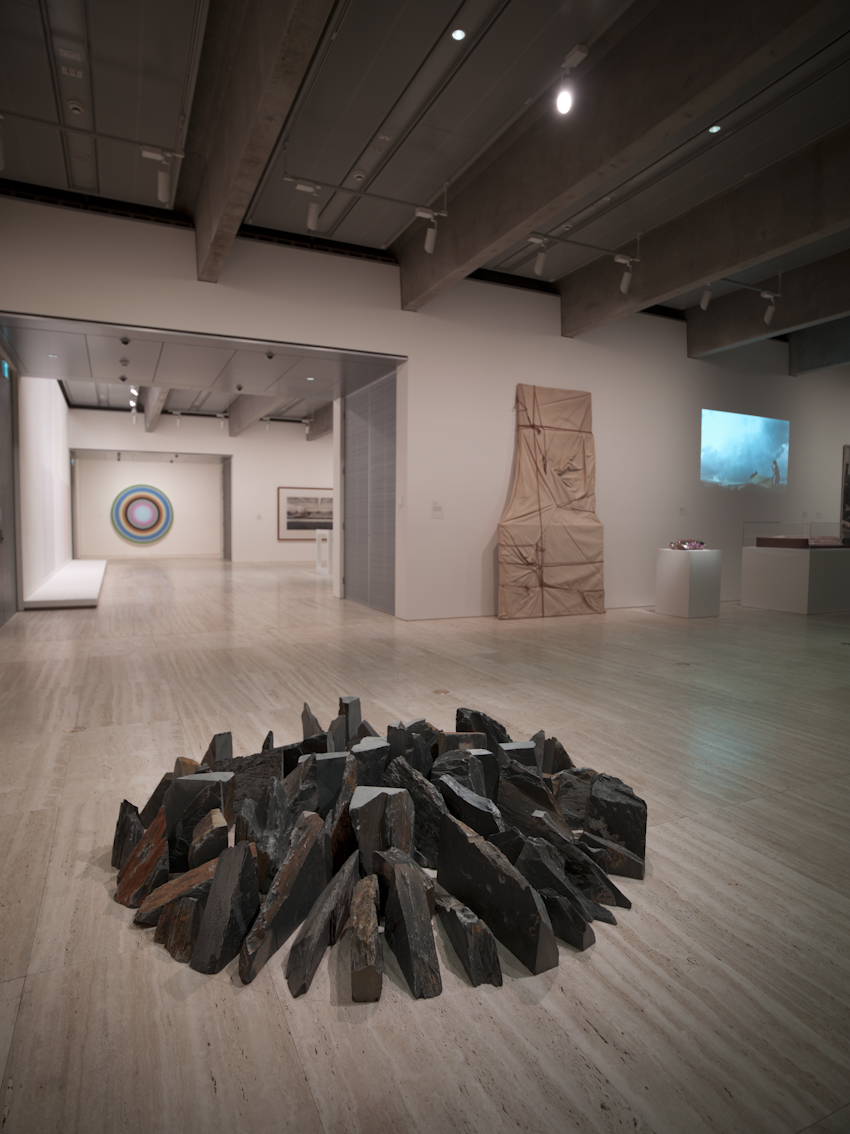 Gallery interior displaying Richard Long's 'Spring Showers Circle', a circular arrangement of dark wooden pieces on a wooden floor, with other contemporary artworks visible in the background.