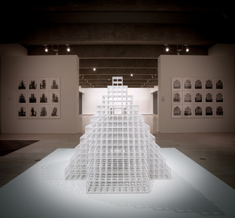 Sol LeWitt's white latticed pyramid sculpture displayed in a gallery with framed artworks on side walls.