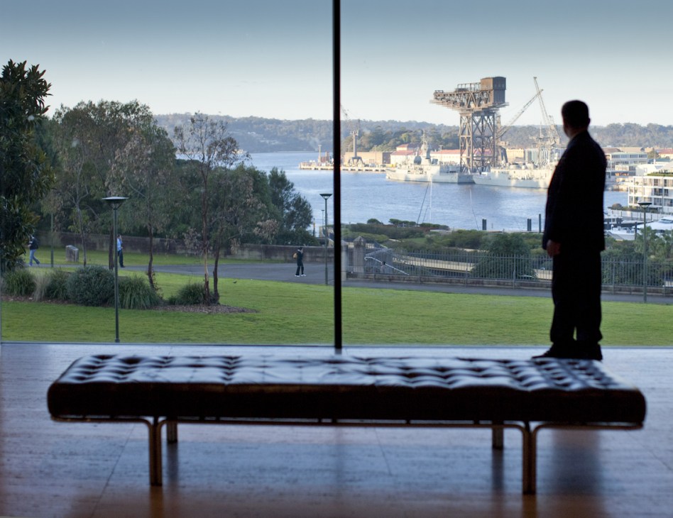 Person standing in gallery viewing Sydney Harbour and parkland through floor-to-ceiling windows